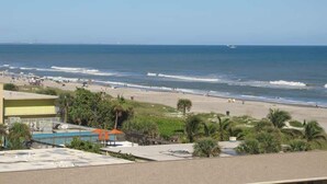 A beach view with a swimming pool, palm trees, and a building in the foreground. - Salt Life Escape: 2B/2B in Oceanfront Complex (Cocoa Beach)