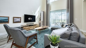 Una sala de estar moderna con un televisor de pantalla plana, sofás grises, una mesa de centro de madera y un ventanal con cortinas.
