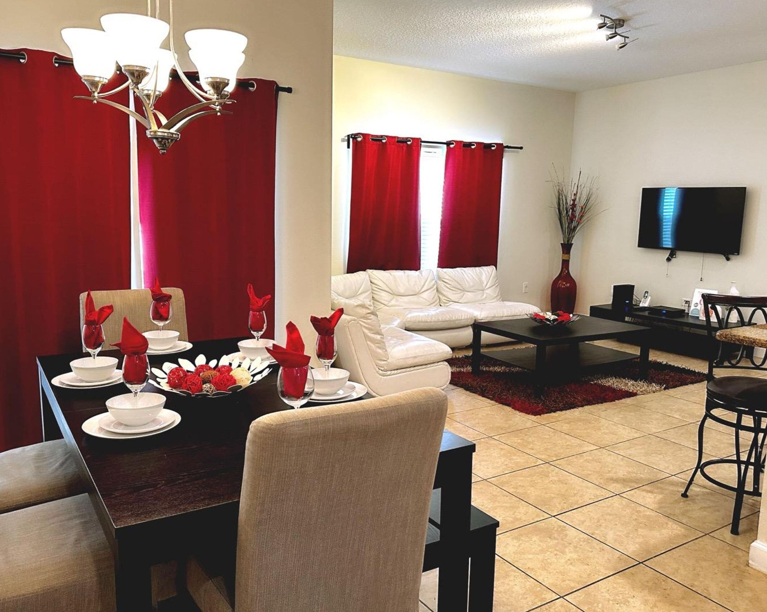 A living room with a dining area, red curtains, a flat-screen TV, and a chandelier.