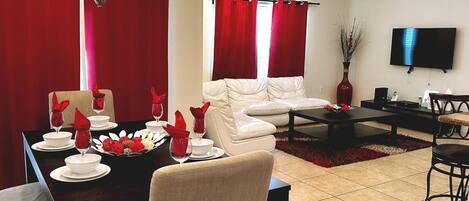 A living room with a dining area, red curtains, a flat-screen TV, and a chandelier.