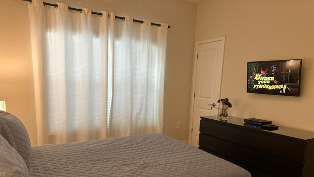 A bedroom with a bed, a dresser, a TV on the wall, and a window with curtains. - 4bed Home in Storey Lake With Private Pool (Kissimmee)