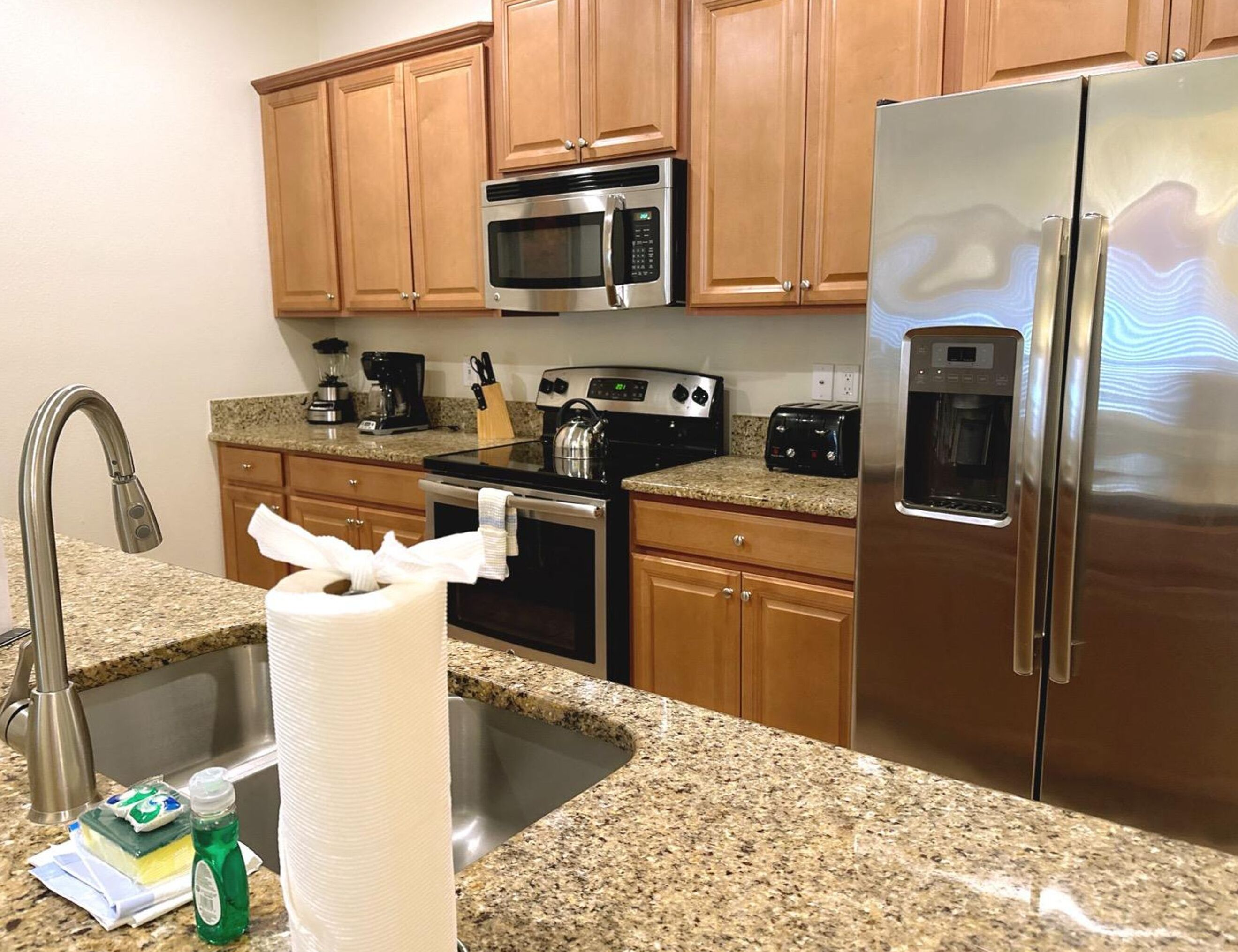 A kitchen with wooden cabinets, stainless steel appliances including a microwave, stove, and refrigerator, a granite countertop, and a sink with a faucet.