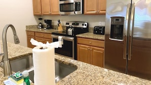 A kitchen with wooden cabinets, stainless steel appliances including a microwave, stove, and refrigerator, a granite countertop, and a sink with a faucet. - 4bed Home in Storey Lake With Private Pool (Kissimmee)