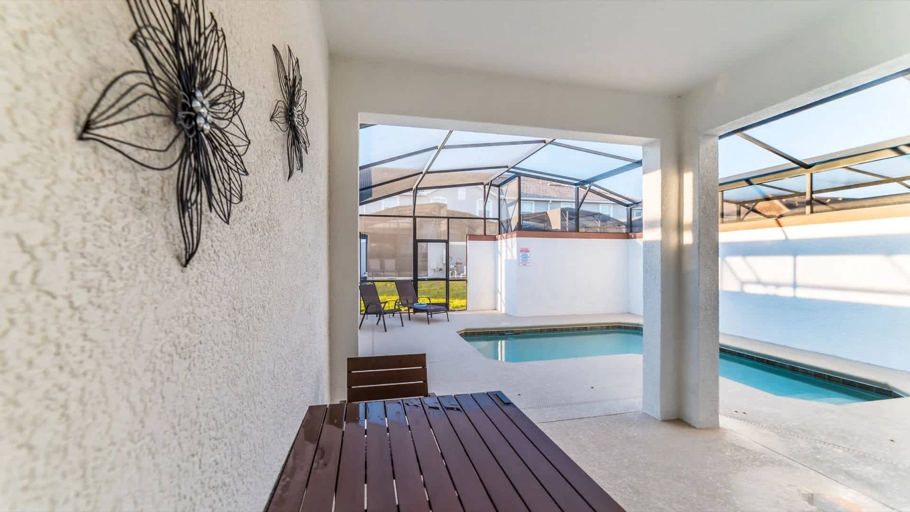 A covered outdoor area with a pool, lounge chairs, and a wooden table.