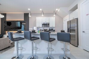 A modern kitchen with white cabinets, a stainless steel refrigerator, and a flat-screen TV mounted above the counter. - Luxury 3BR Home With Mario Bros Room Near Disney (Kissimmee)