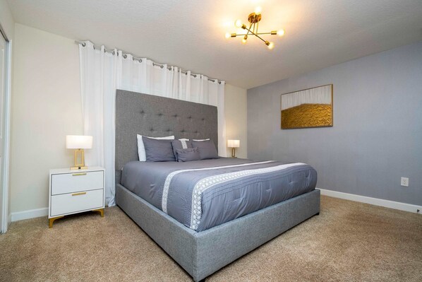 Une chambre à coucher avec un lit recouvert d’un tissu gris, des tables de chevet blanches, une tête de lit grise et un luminaire au plafond.