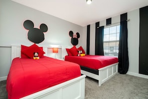 Habitación de hotel con dos camas, cada una decorada con la cabeza de Mickey Mouse, ropa de cama roja y cabecero blanco.