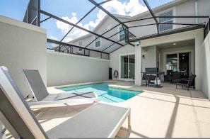 Zona de piscina con sillas de descanso, una mesa y un porche cubierto.