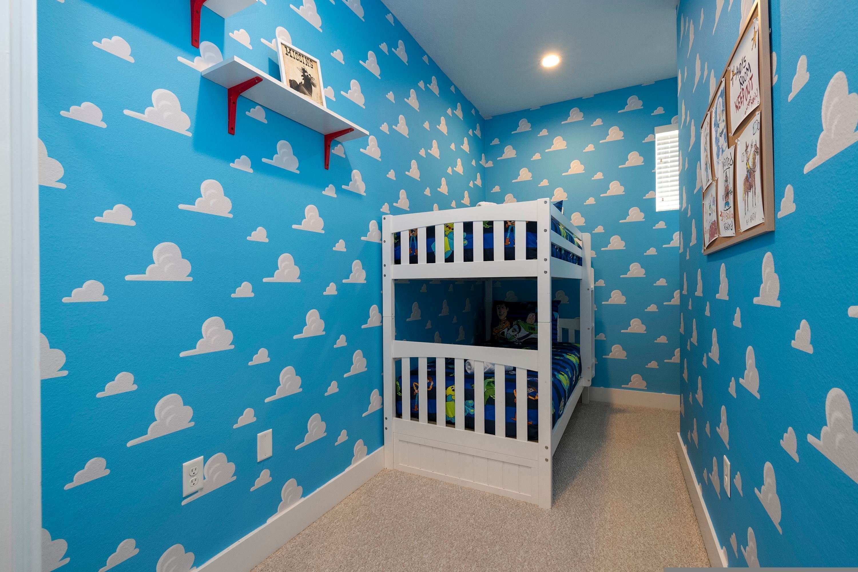 Een kamer met een stapelbed, blauwe wanden met wolkenpatronen, een plankje met een foto en een raam.
