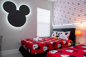 Ein Zimmer mit zwei Betten, eines mit Bettwäsche mit Mickey Mouse Motiv und das andere mit Bettwäsche mit Minnie Mouse Motiv. An der Wand ist eine große Silhouette von Mickey Mouse zu sehen, ein Fenster mit Rollläden und die Decke ist mit Mickey Mouse Tapete bedruckt.