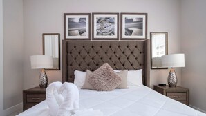A neatly made bed with a tufted headboard, two bedside tables with lamps, and three framed pictures on the wall. - Modern Chic Home Mickey and Toy Story Rooms (Kissimmee)