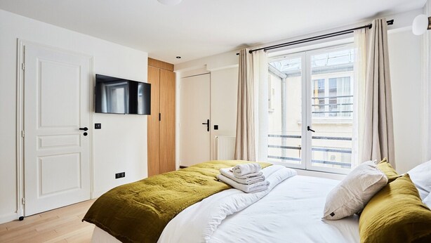 A bedroom with a bed, a television mounted on the wall, a window with curtains, and a door. - Chic Studio AC - Center of Paris-bourse (Paris)