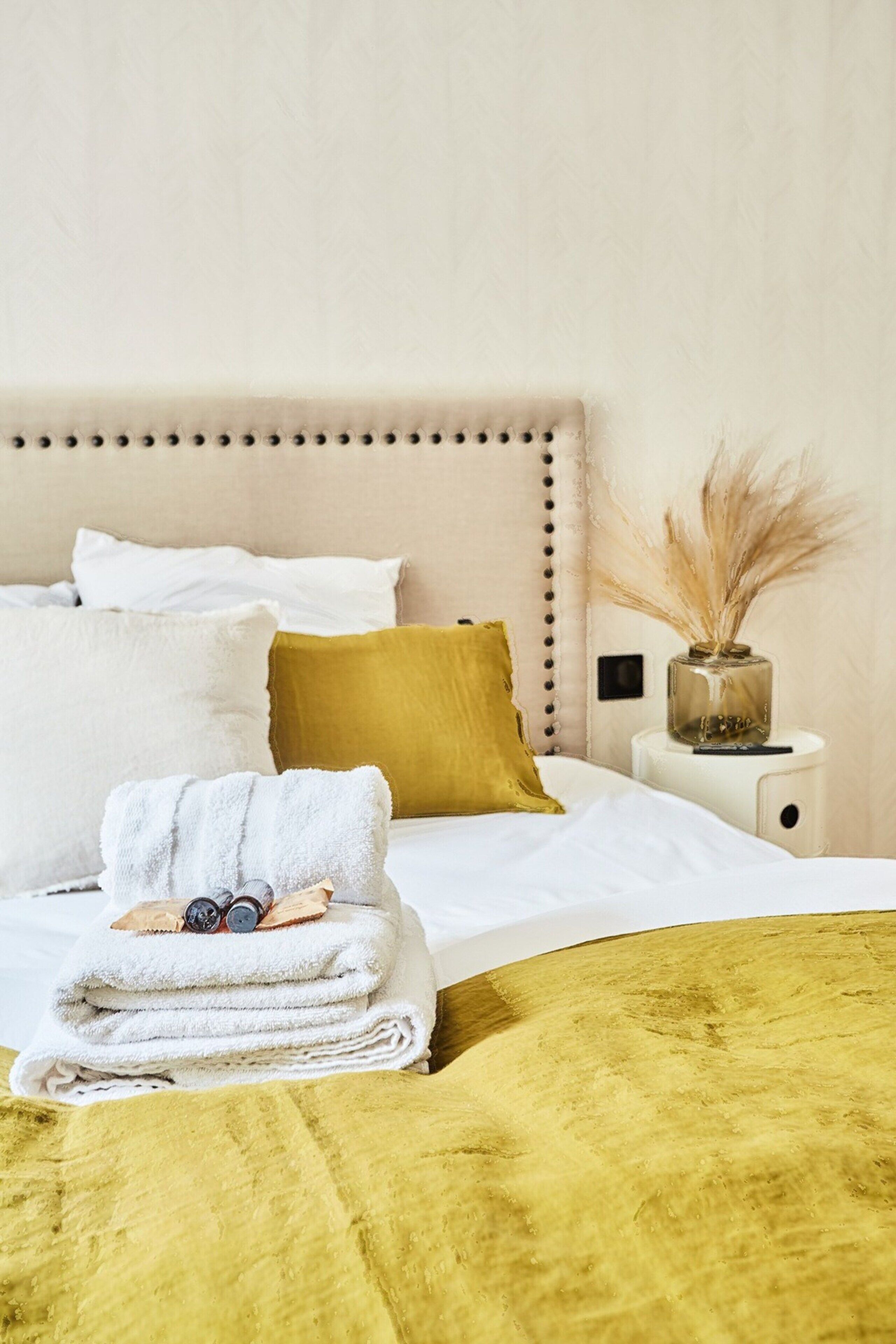Una cama bien tendida con almohadas blancas y amarillas, una mesita de noche con un florero, y una manta amarilla.