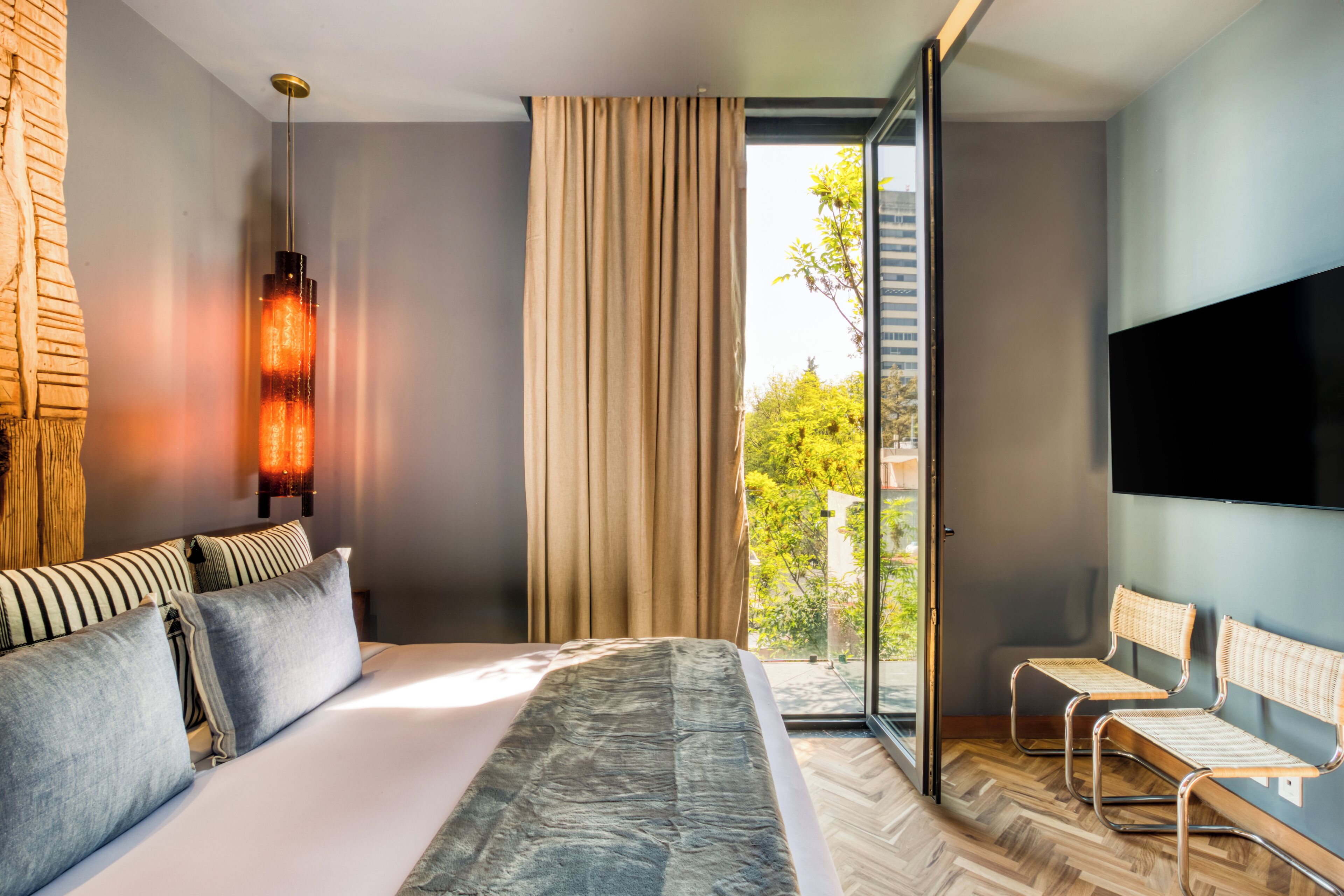 A modern hotel room with a large bed, a TV, a window with curtains, and a view of greenery outside.