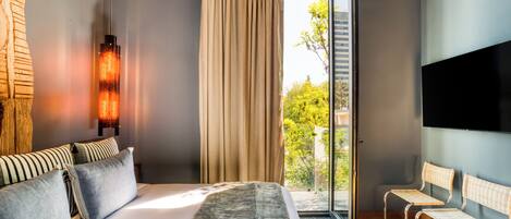 A modern hotel room with a large bed, a TV, a window with curtains, and a view of greenery outside.