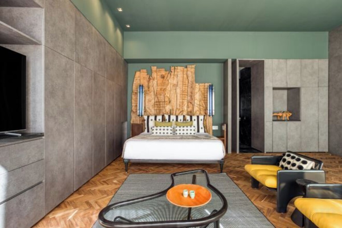 A modern hotel room with a large bed, a flat-screen TV, a seating area with a table, and a decorative wall piece.