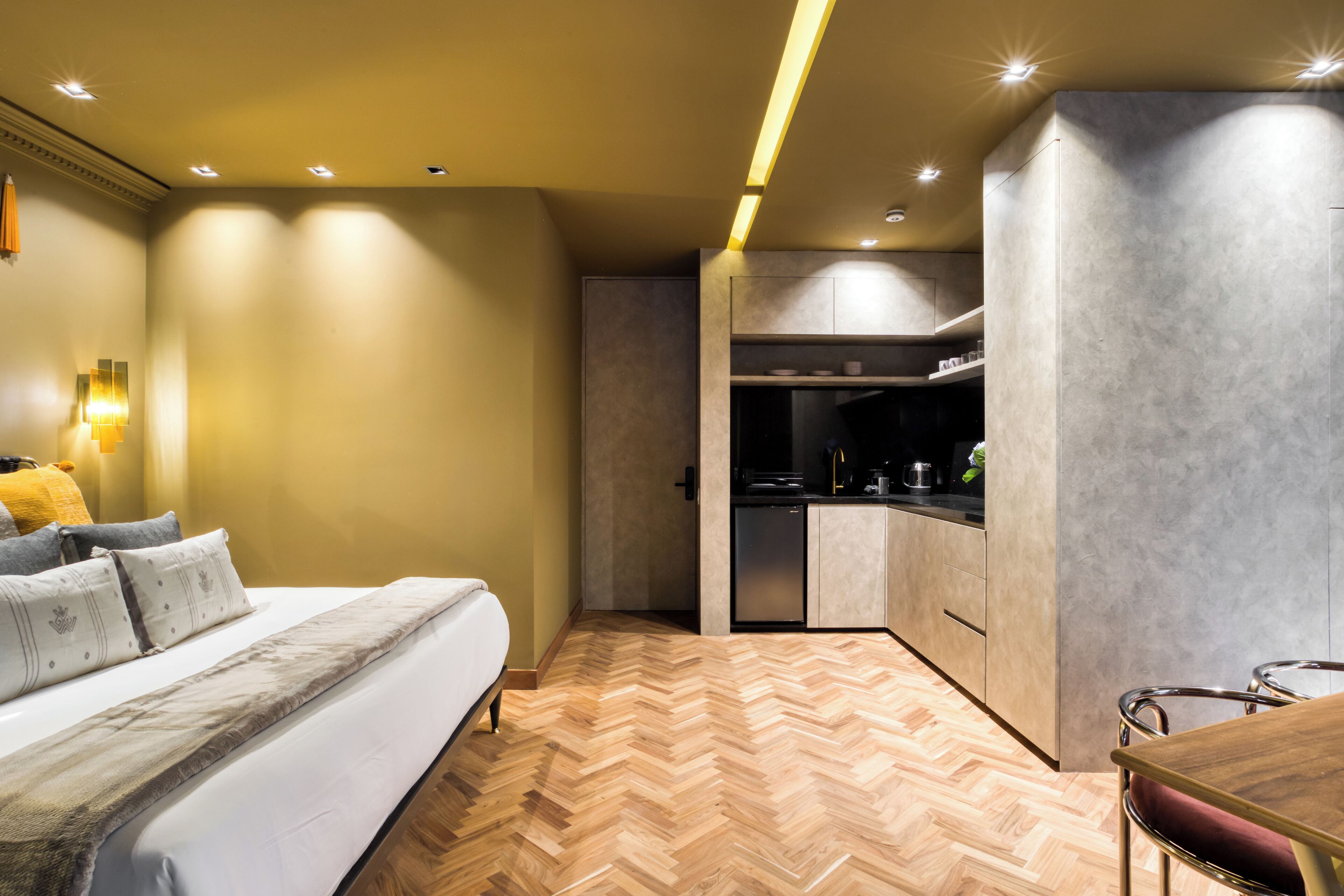 A modern hotel room with a bed, a wooden herringbone floor, a kitchenette, and a dining area with a table and chairs.