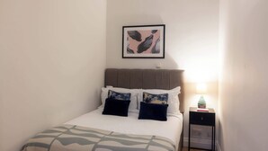 A neatly made bed with a patterned bedspread, a headboard, and two pillows. There is a framed picture on the wall above the bed and a small bedside table with a lamp and a book.