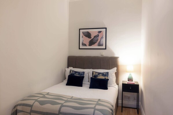 A neatly made bed with a patterned bedspread, a headboard, and two pillows. There is a framed picture on the wall above the bed and a small bedside table with a lamp and a book.