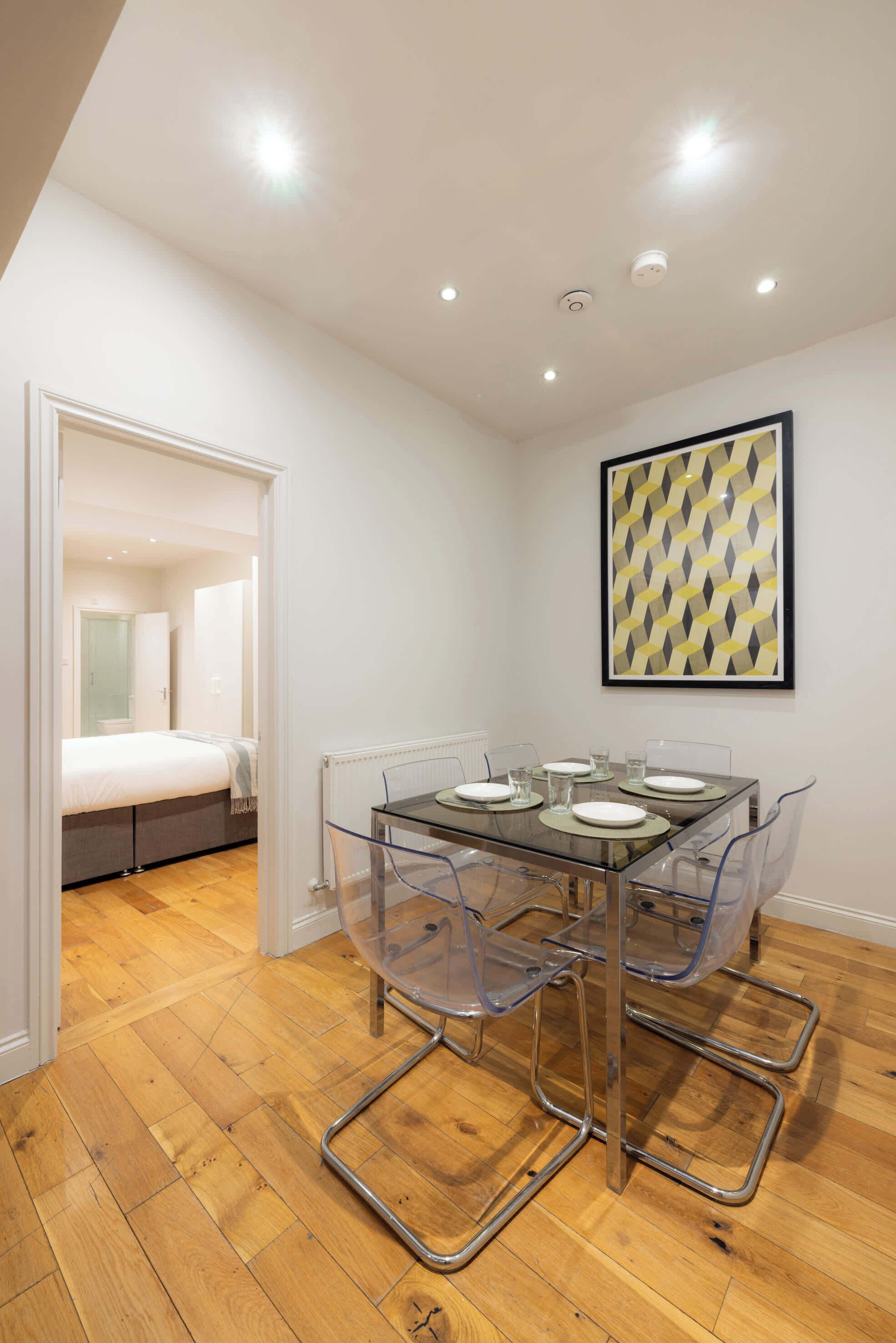 A modern room with a glass dining table, clear chairs, a bed, and a framed geometric artwork on the wall.