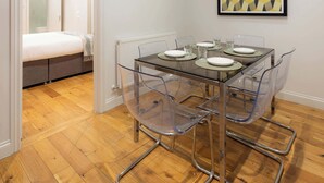 Una habitación moderna con una mesa de comedor de vidrio, sillas transparentes, una cama y una obra de arte geométrica enmarcada en la pared.