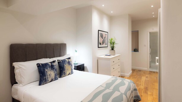 Un dormitorio con cama, mesita de noche, cómoda y baño visible a través de una puerta abierta.