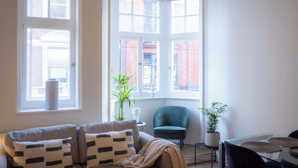 Una habitación luminosa con un ventanal, un sofá con cojines estampados, un sillón color turquesa y una mesa de comedor con individuales.