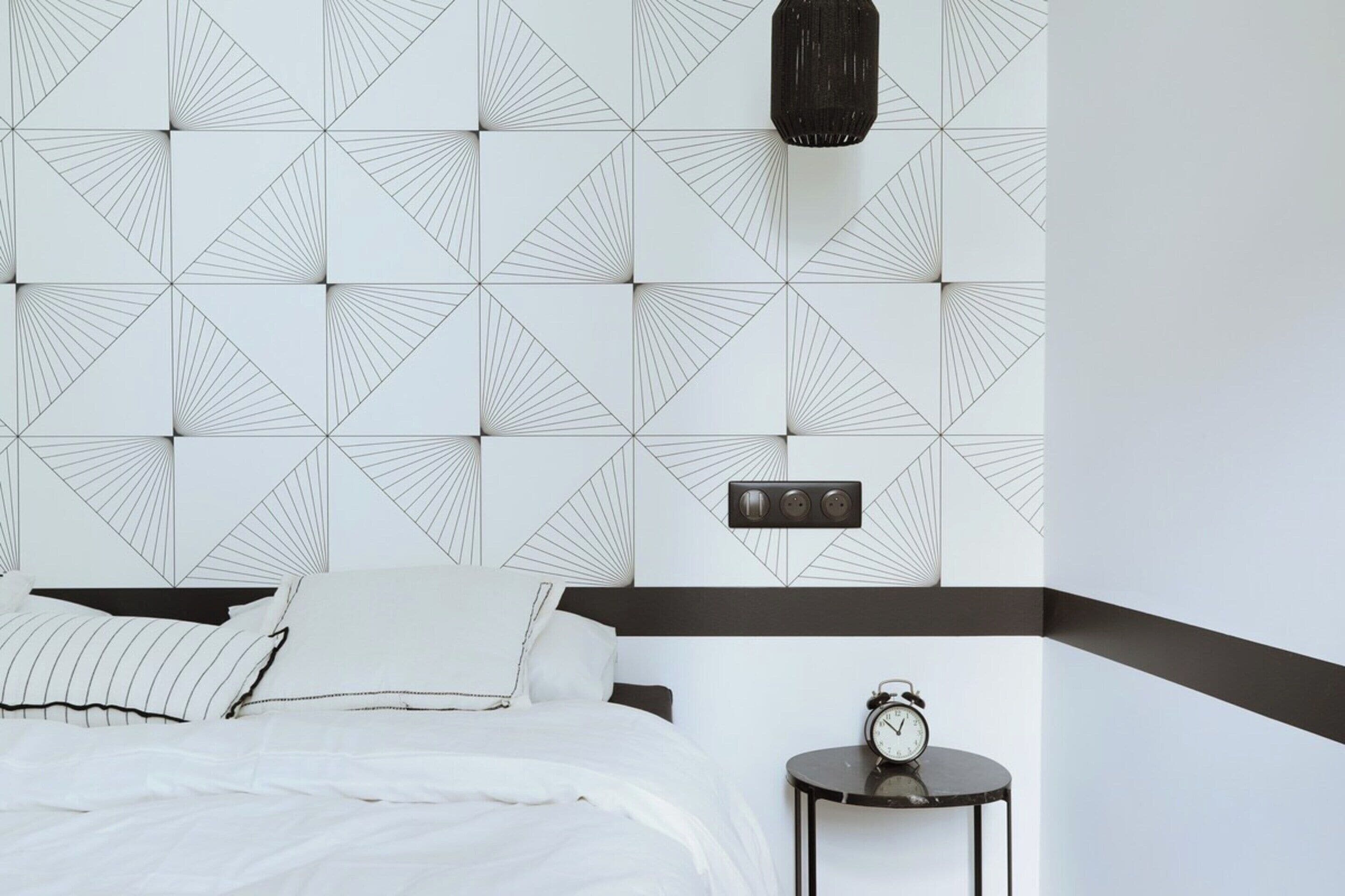 Une chambre d’hôtel avec une tête de lit à motifs géométriques, un lit avec une literie blanche, une petite table de chevet avec une horloge et un mur d’accent noir.