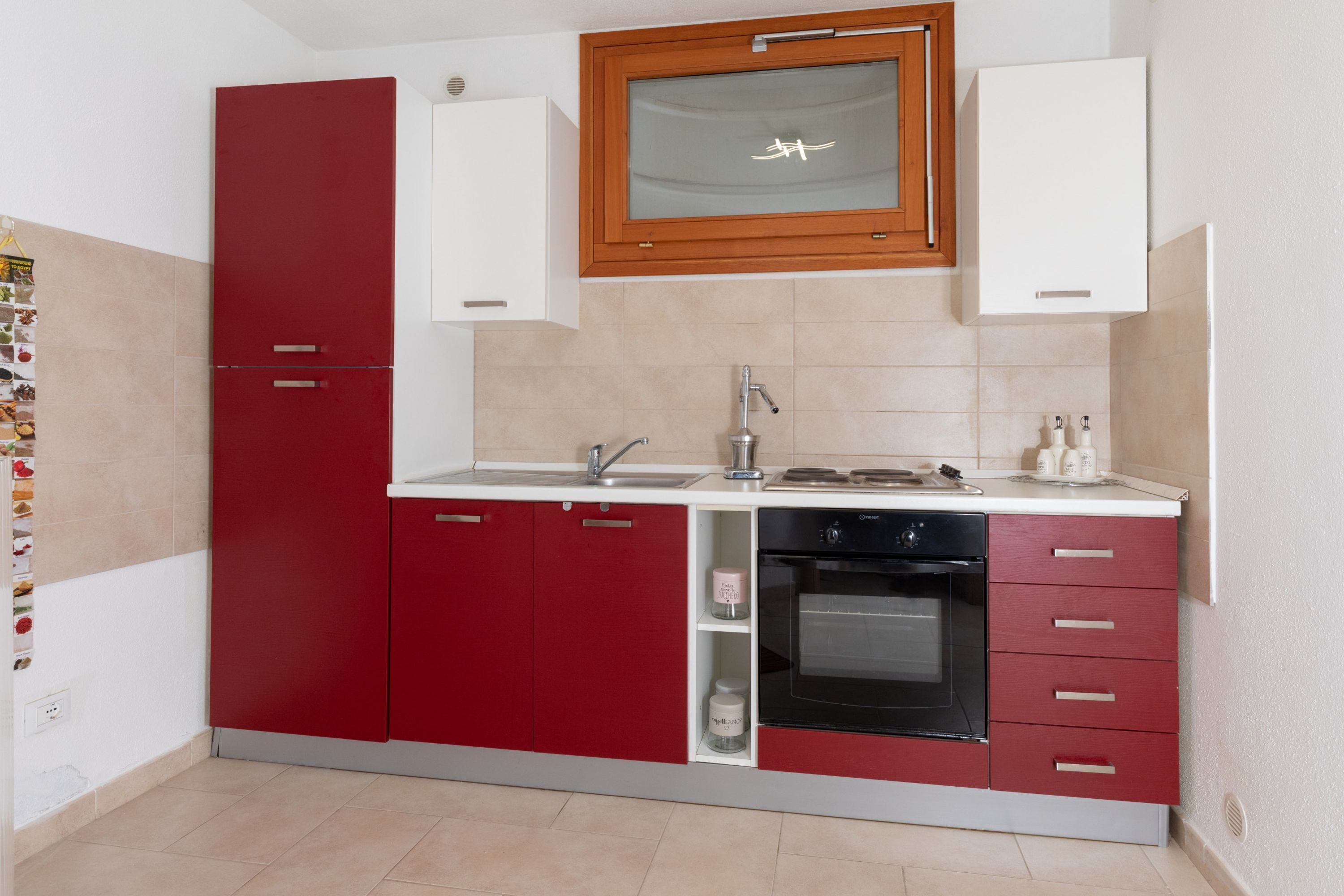 A compact kitchen with a red and white color scheme, featuring a built-in oven, a sink, and a window above the countertop.