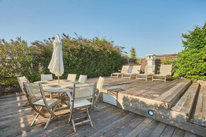 Um deck externo com uma mesa e cadeiras de madeira, cercado por vegetação.