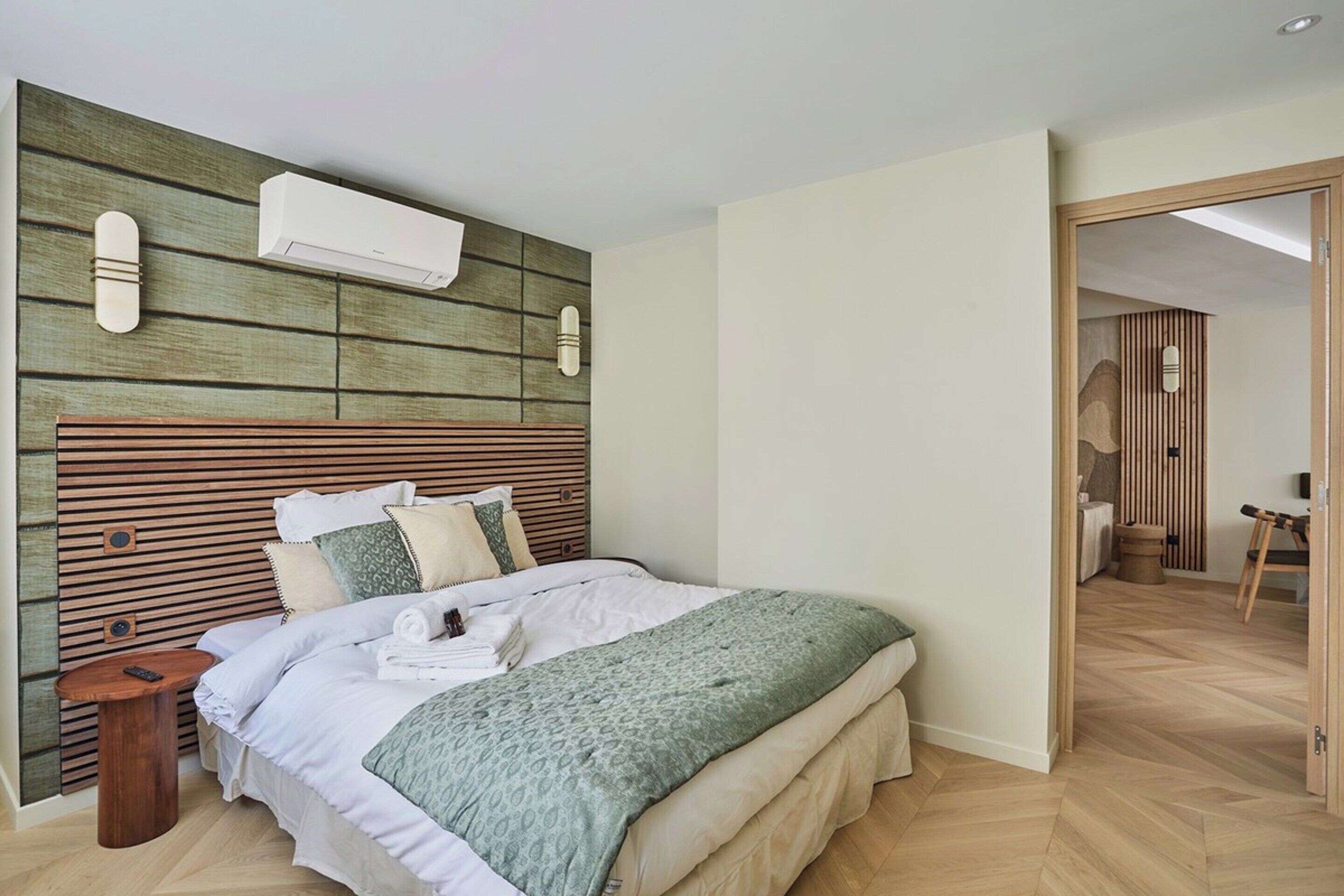 A modern bedroom with a wooden headboard, a bed with white linens, a green throw blanket, and a small bedside table with a remote control.