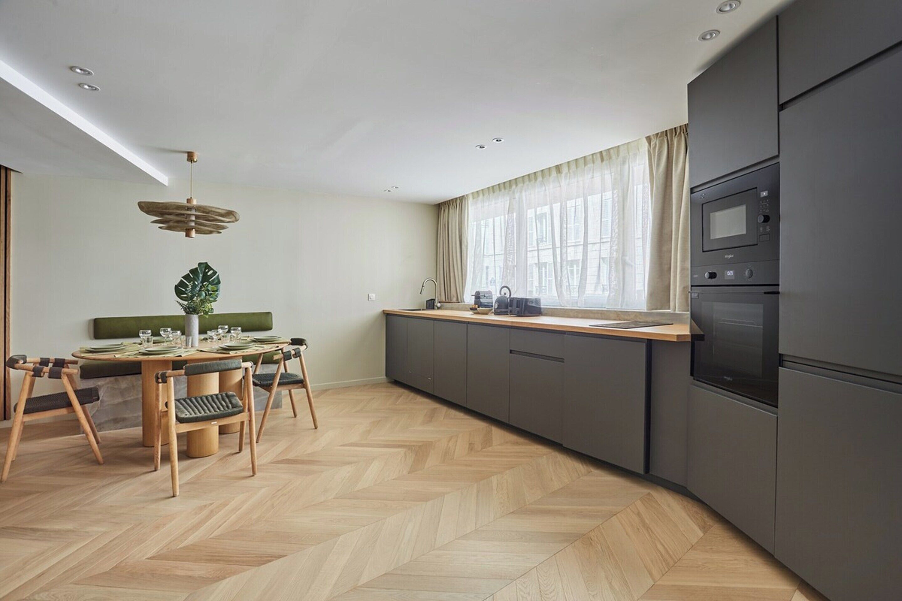 A modern kitchen with a herringbone wood floor, a dining area with a wooden table and chairs, and a large window with sheer curtains.
