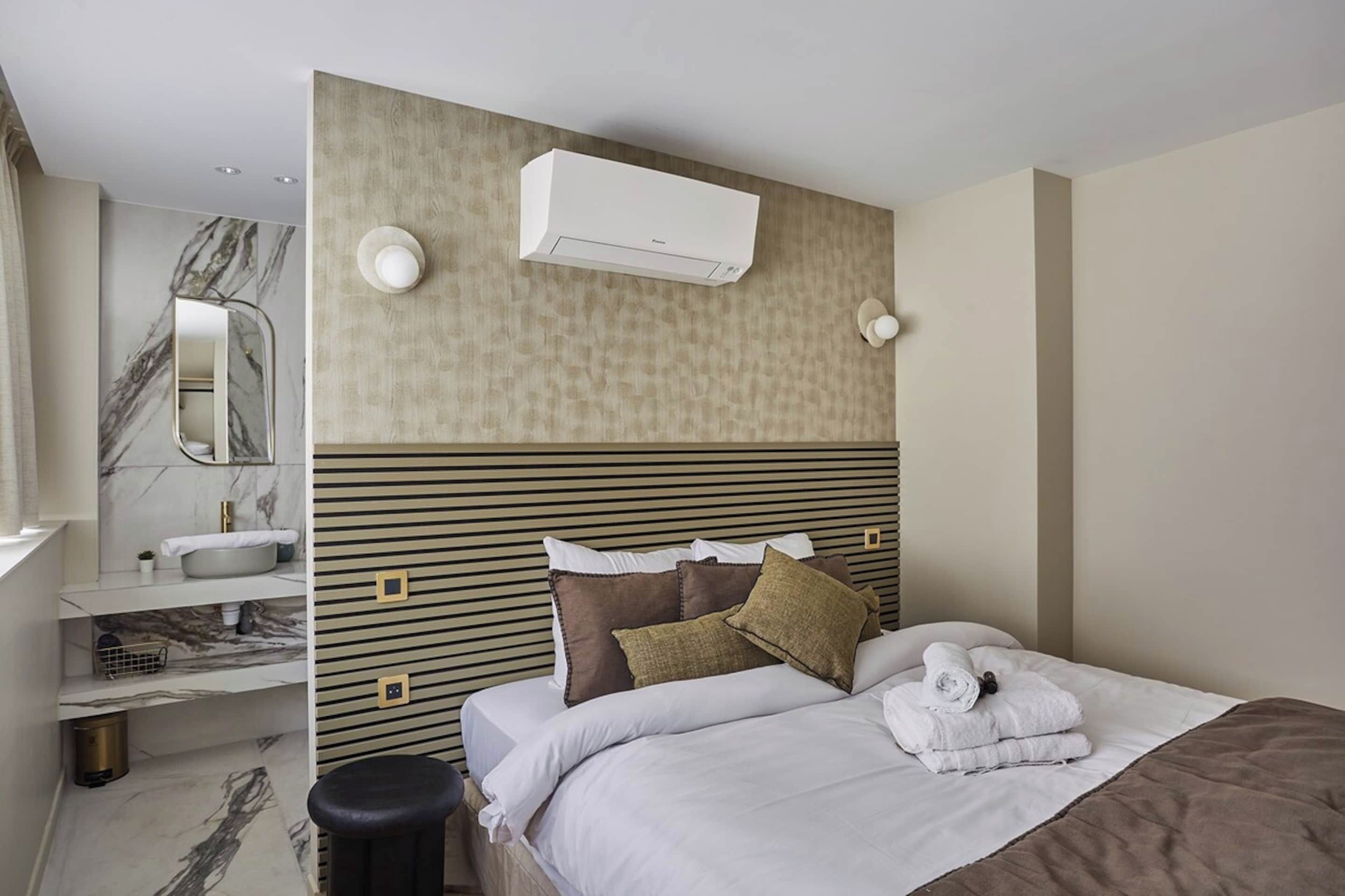 A hotel room with a marble bathroom, a bed with pillows, a bedside table, and a wall-mounted air conditioner.
