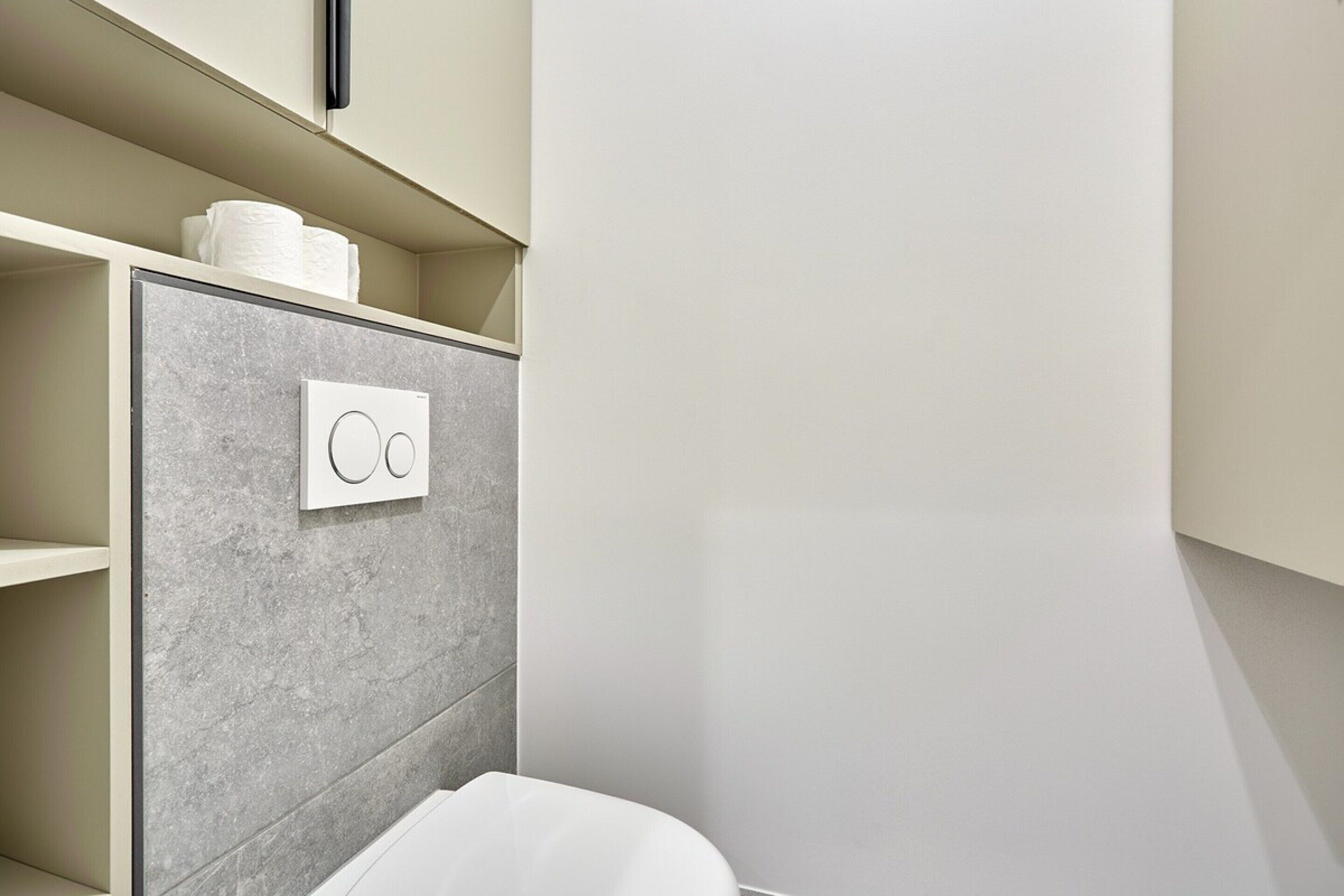 A modern bathroom with a grey tiled wall, a toilet, and a shelf with toiletries.
