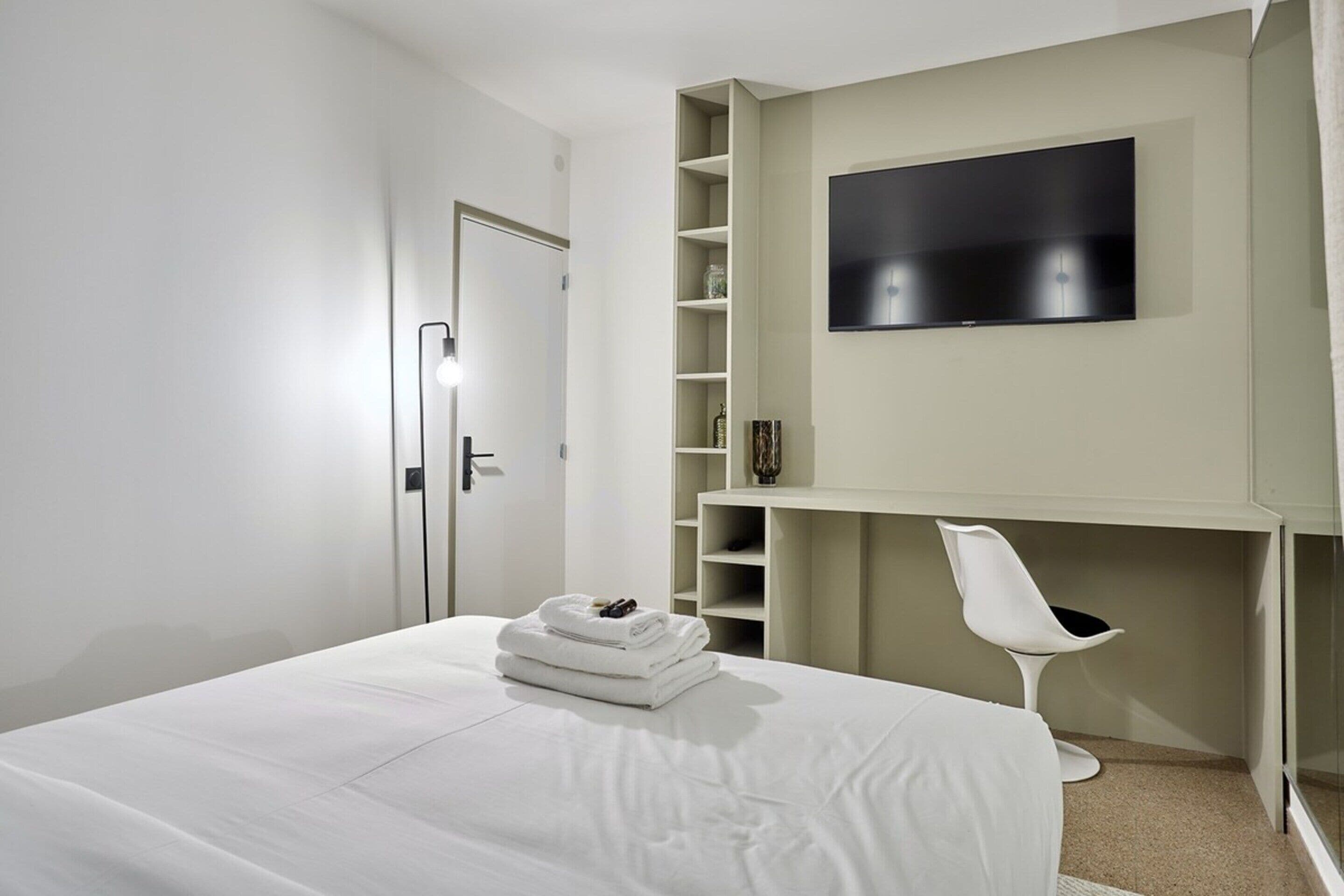 A modern bedroom with a bed, a TV mounted on the wall, and a white chair.