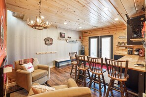 A cozy cabin interior with a wooden ceiling, a dining area with wooden chairs, a sofa with a plaid throw, and a coffee station.