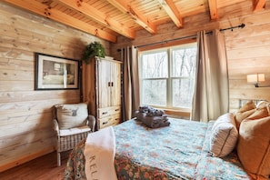 Una habitación de cabaña acogedora, con paredes de madera, una cama con ropa de cama floral, una silla de mimbre y una ventana con cortinas.