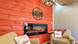 A cozy room with a red wooden wall, a fireplace, two beige armchairs, and a 'Home' pillow on one armchair.
