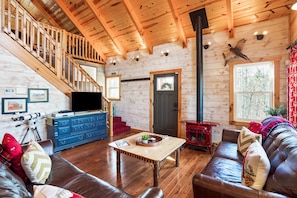 Interior acogedor de una cabaña con escalera de madera, chimenea, televisor y sofá de cuero.