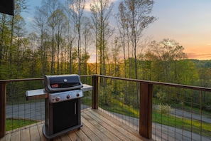 A barbecue grill on a wooden deck with a view of a forested area and a road in the background.