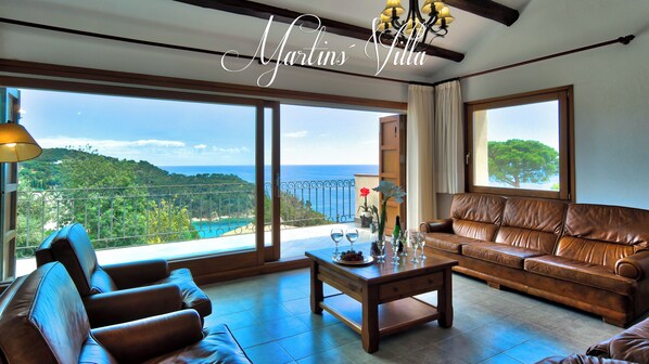 A spacious living room with a leather sofa, a wooden coffee table, and a view of the sea through large windows.