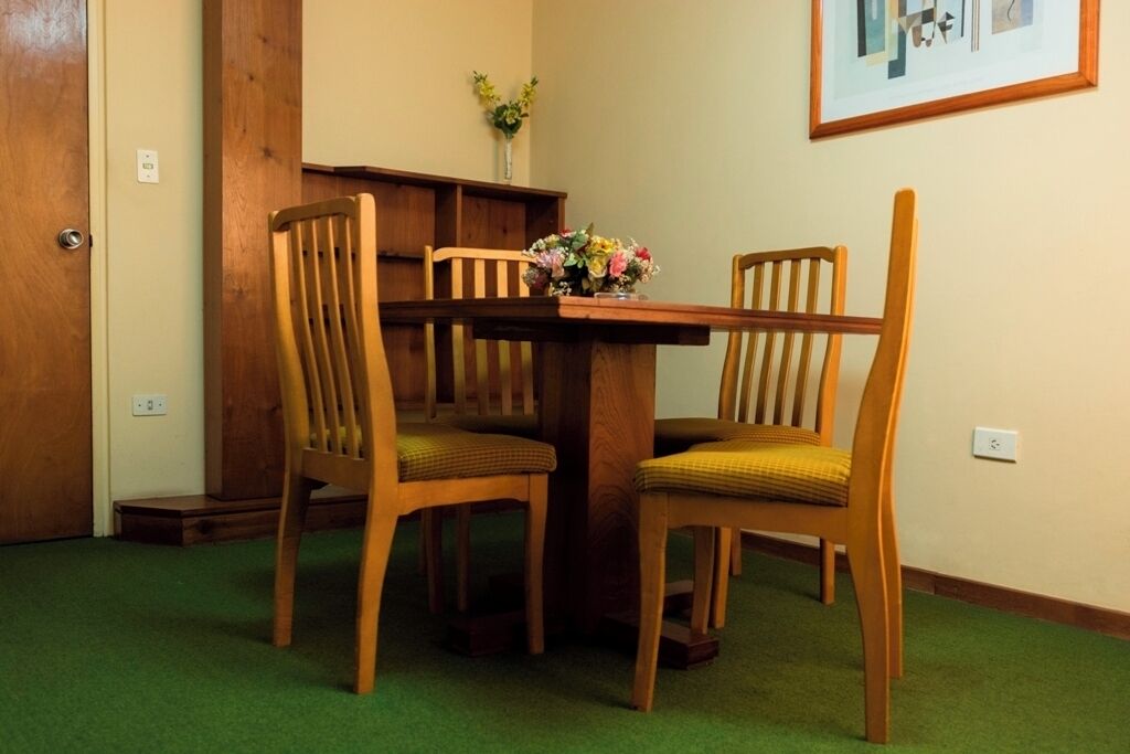 A dining area with a wooden table and two chairs, a vase with flowers on the table, and a framed picture on the wall.
