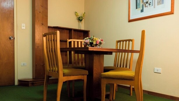 A dining area with a wooden table and two chairs, a vase with flowers on the table, and a framed picture on the wall. - Hotel del Jardín (San Miguel de Tucuman)