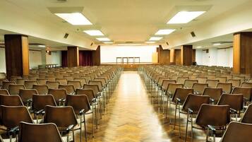 Un salón de conferencias amplio y bien iluminado, con filas de sillas mirando hacia un escenario.