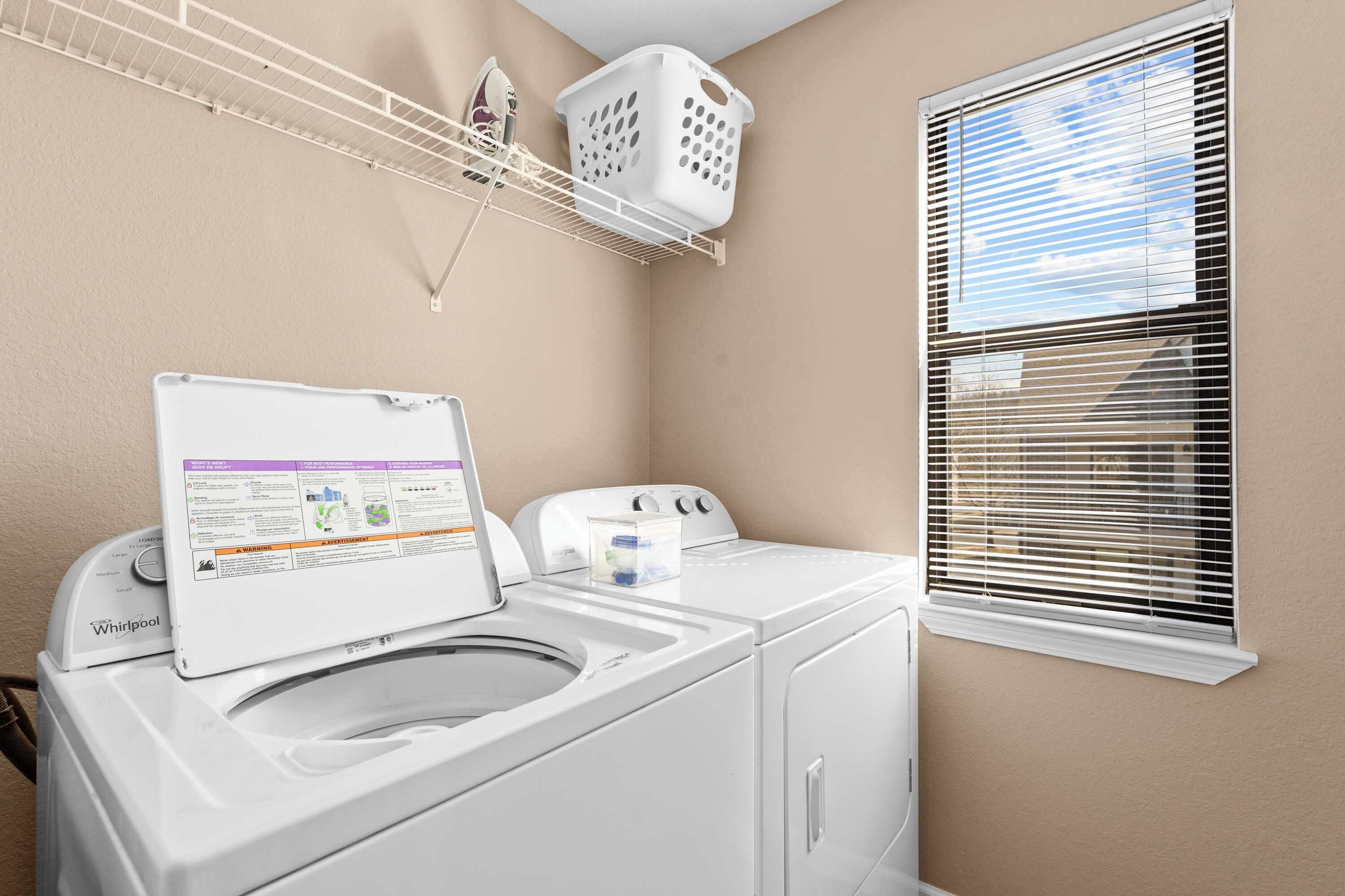 A laundry room with a Whirlpool washer and dryer, a drying rack, a laundry basket, and a window with blinds.