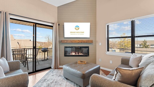 A modern living room with a fireplace, sliding glass doors to a balcony, and a view of the neighborhood. - Branson Blessing Condo for 8! (Branson)