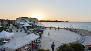A coastal promenade with market stalls, a building with a white facade, and a sunset over the sea.