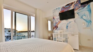 A hotel room with a large bed, a desk, a flat-screen TV mounted on the wall, and a view of the sea through large windows.