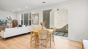 Een moderne woonkamer met een witte bank, een eettafel met rieten stoelen, en een groot raam met uitzicht op een tuin.