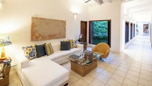 A spacious living room with a white sofa, a glass coffee table, and a yellow armchair. There is a ceiling fan, a large abstract painting, and a view of greenery through a window. - Yeguas V43 by Wynwood House (Anapoima)